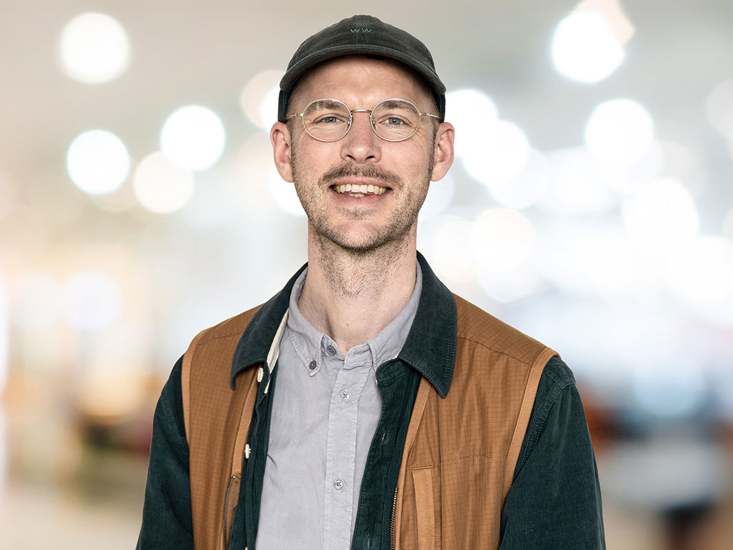Portrait of a smiling man looking in to the camera. He wears a cap, silver colored glases and a brown vest over a shirt.