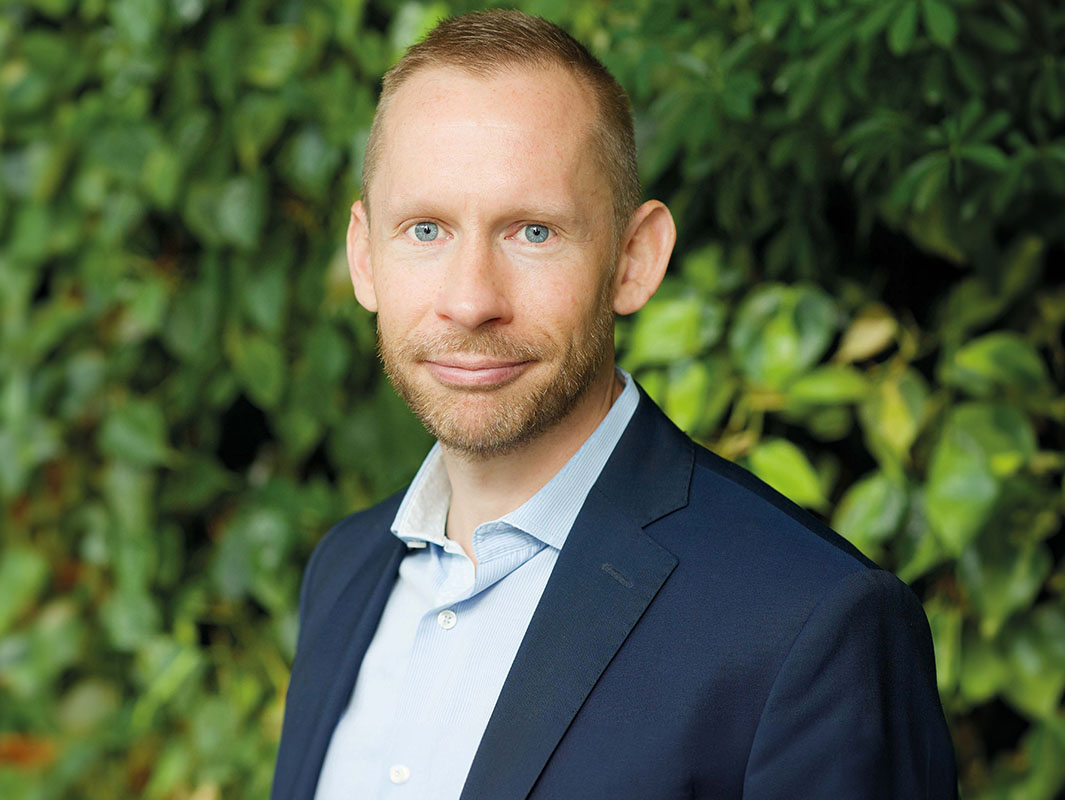 Portrait of a smiling man looking in to the camera. He has blonde hair and wears a dark blue blazer jacket.