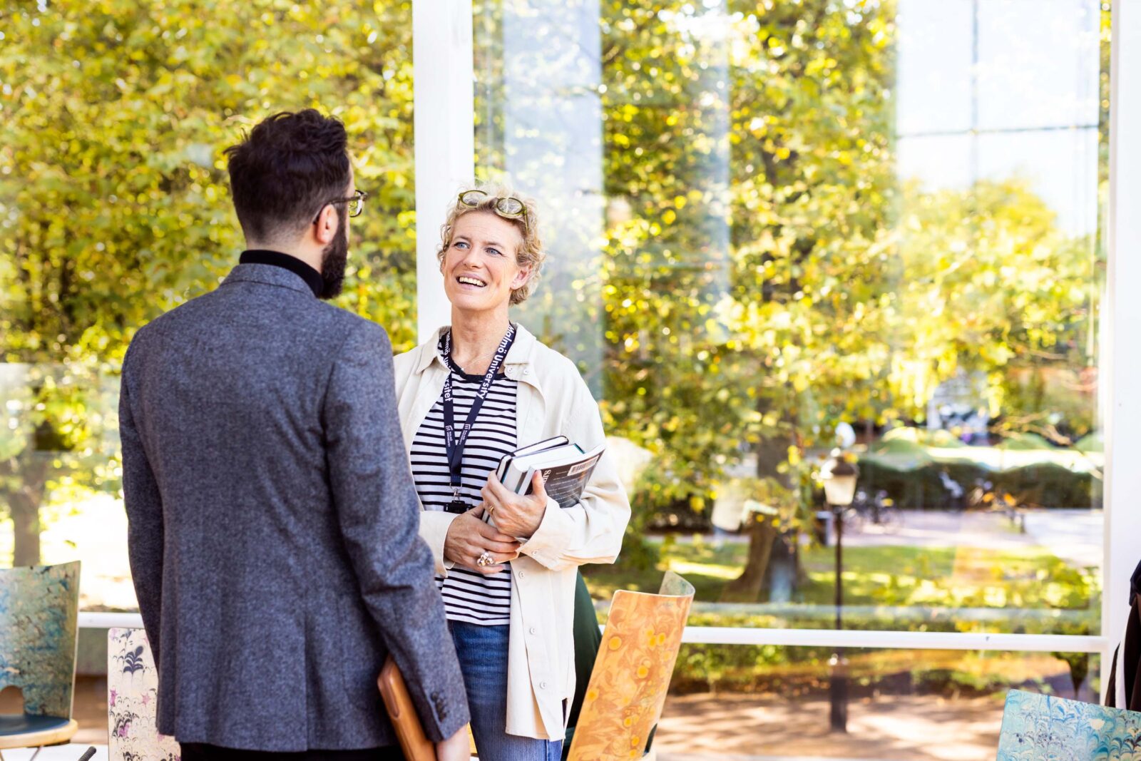Two colleagues talking to each other. The male collegue is wearing a blur blazer and the women is wearing a white blaxer over a striped shirt.